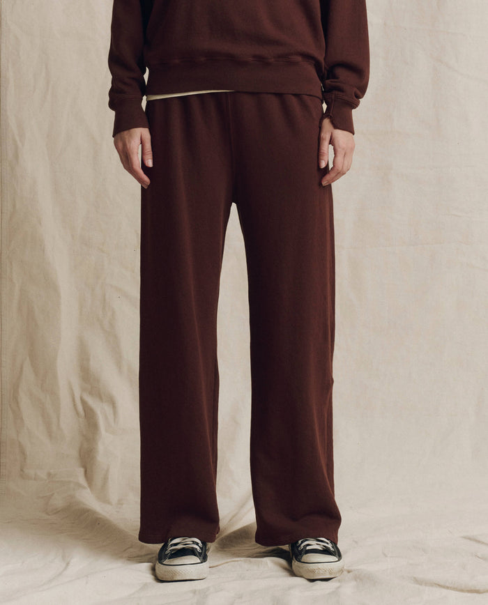 Wearing The Lap Sweatpant in Solid Driftwood by THE GREAT. F26 SEASONAL CORE—a loose, cropped straight-leg brown pant—paired with a coordinating brown long-sleeve top and black-and-white sneakers, the person stands against a light fabric backdrop.