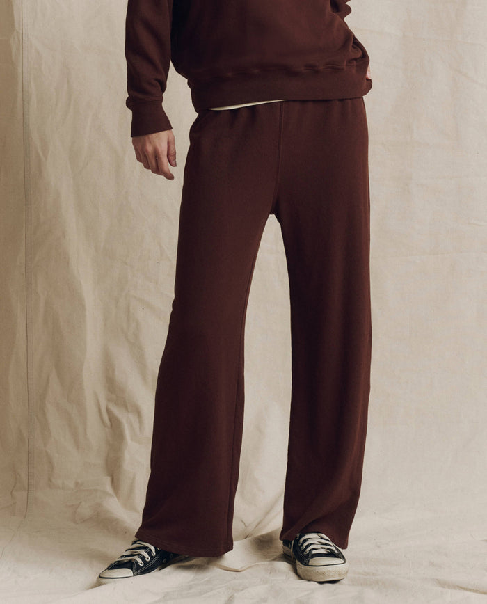 A person wearing the loose-fitting The Lap Sweatpant in Driftwood by THE GREAT. F26 SEASONAL CORE, featuring a cropped straight leg, paired with a matching top and black sneakers, stands on a light fabric backdrop. Only the lower half of their body is visible.