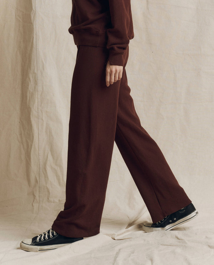 A person stands in front of a beige fabric backdrop wearing The Lap Sweatpant. Solid -- Driftwood from THE GREAT. F26 SEASONAL CORE—a pair of cropped, straight leg sweatpants handcrafted in Los Angeles—paired with a matching sweatshirt and black sneakers. The person’s head is not visible in the image.