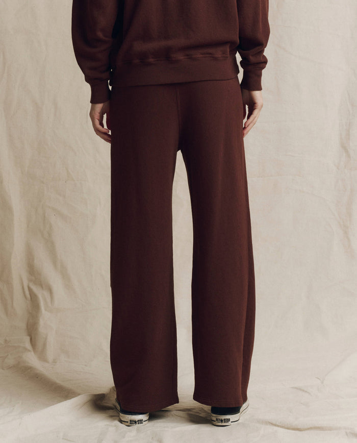 A person stands on a neutral background wearing THE GREAT. F26 SEASONAL CORE The Lap Sweatpant, Solid -- Driftwood, paired with a matching brown top and white sneakers; only the lower half of the body is visible.