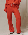 Person in a handcrafted Los Angeles orange sweatshirt and THE GREAT. SP26 SEASONAL CORE The Lap Sweatpant in Persimmon with white drawstring, standing on a light-colored floor, partially visible white sneakers.