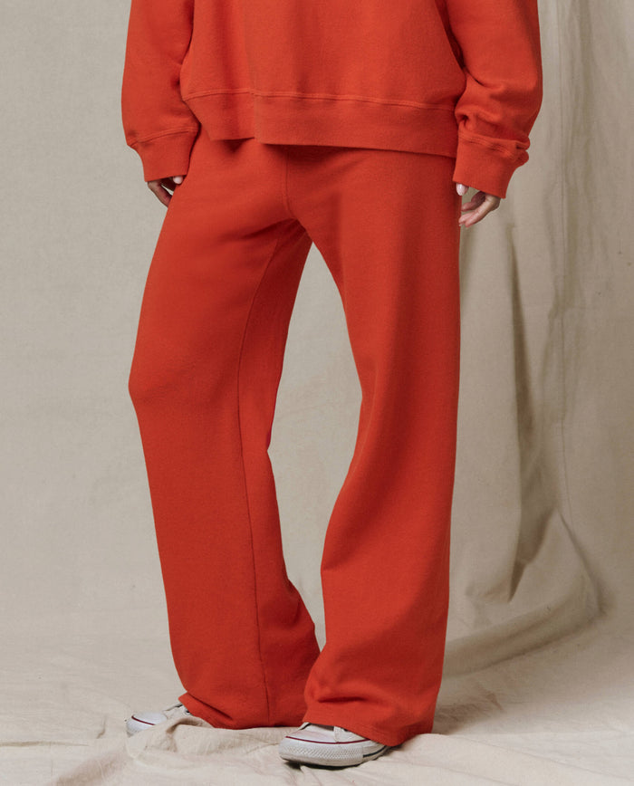 Person wearing THE GREAT. SP26 SEASONAL CORE The Lap Sweatpant in Persimmon—handcrafted, loose-fitting, cropped straight leg—styled with a matching orange sweatshirt and white sneakers, standing against a plain beige background.