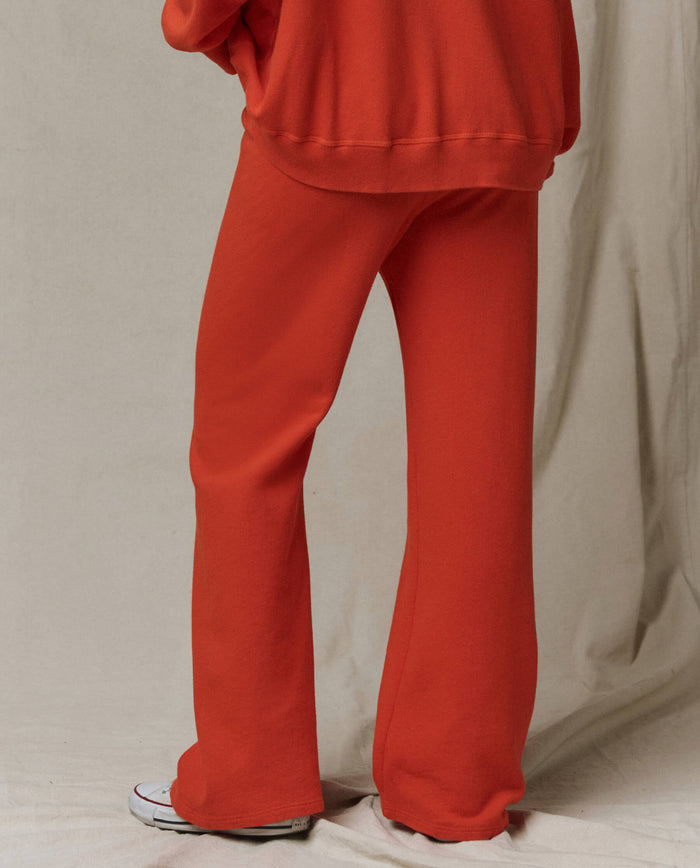 Model wears THE GREAT. SP26 SEASONAL CORE The Lap Sweatpant in Persimmon, a loose-fitting red pant with cropped straight leg, paired with a matching top against a light beige fabric backdrop. Only the lower body is visible.