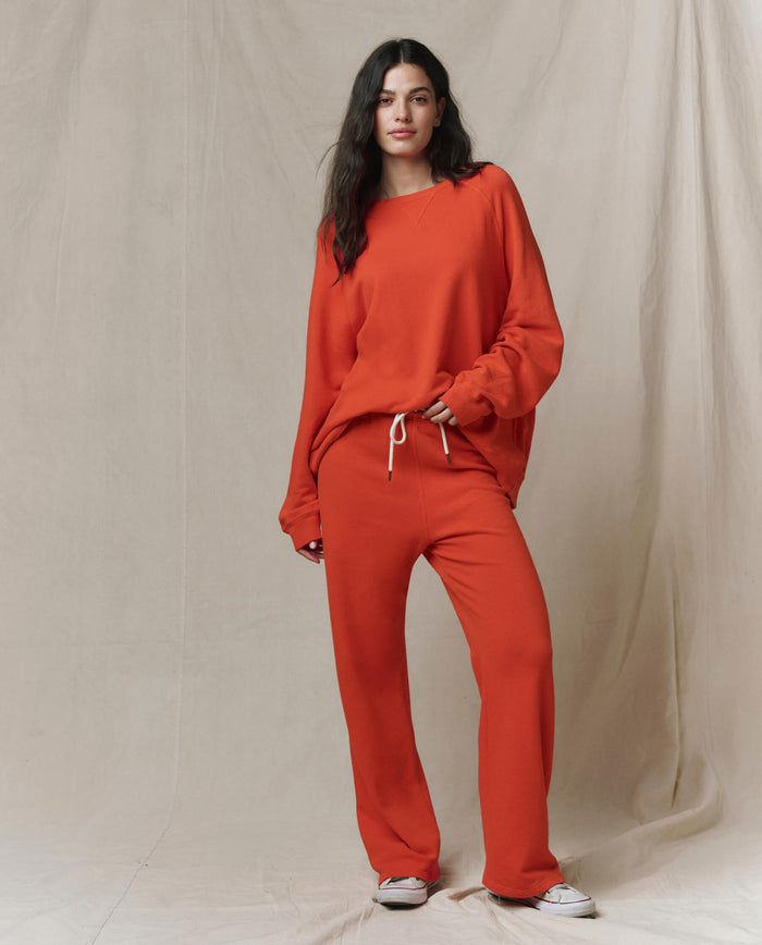 A person stands against a neutral backdrop wearing a loose-fitting, long-sleeve orange top and THE GREAT. SP26 SEASONAL CORE The Lap Sweatpant in Persimmon, featuring a cropped straight leg with white drawstrings and white sneakers.