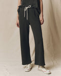 Person wearing loose, washed black The Lap Sweatpant by THE GREAT. CORE KNITS and white high-top sneakers, standing against a beige fabric backdrop.