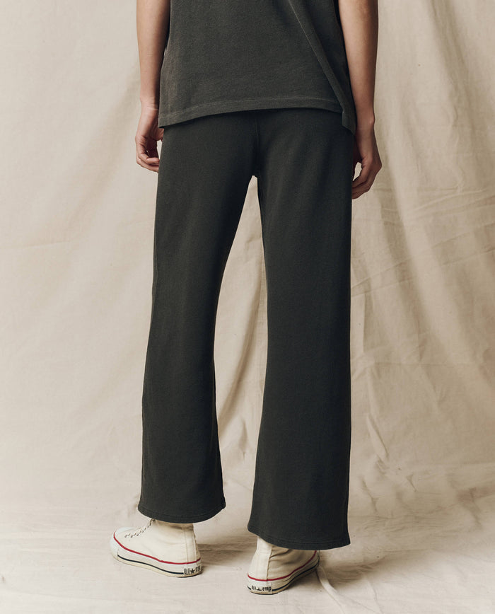 A person stands facing away, wearing THE GREAT. CORE KNITS The Lap Sweatpant. Solid -- Washed Black with a matching dark top and white sneakers, set against a beige fabric background.