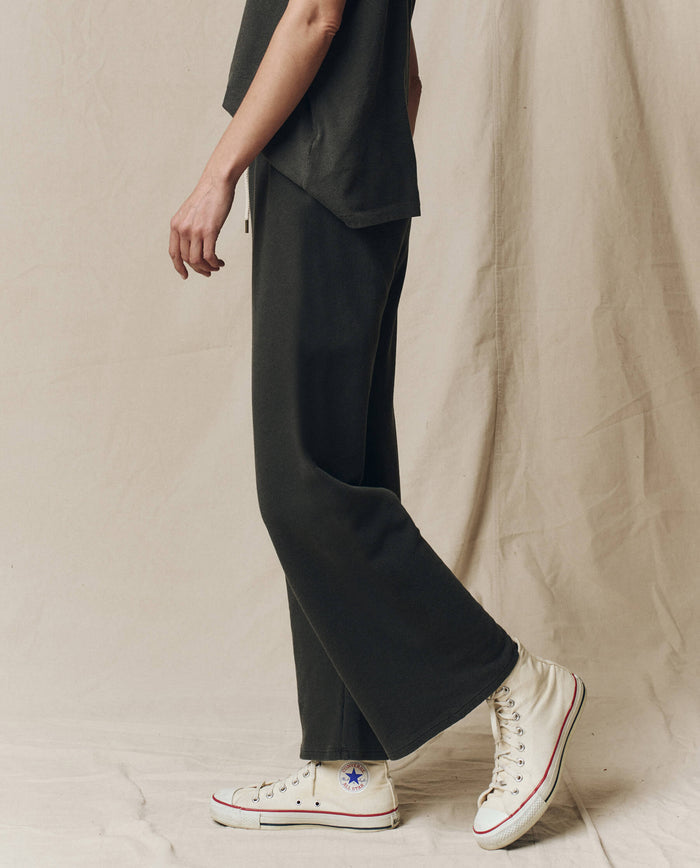 Person wearing THE GREAT. CORE KNITS The Lap Sweatpant in Solid -- Washed Black, a matching short-sleeve top, and white high-top Converse sneakers stands against a neutral fabric backdrop.