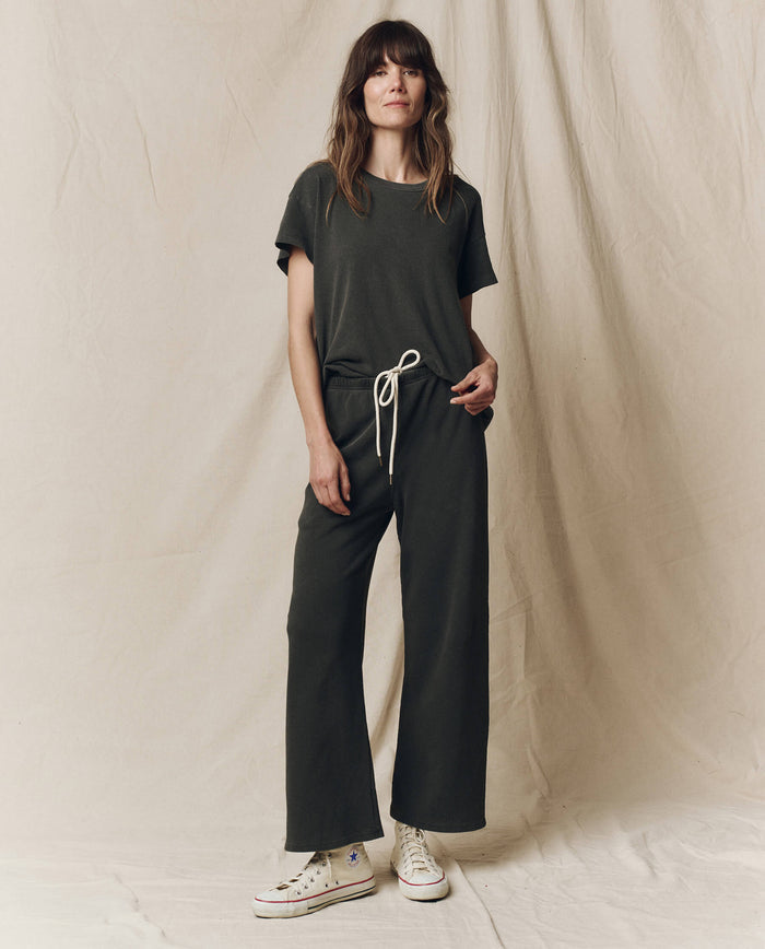 A woman stands against a beige fabric backdrop wearing a dark green short-sleeve top and matching THE GREAT. CORE KNITS The Lap Sweatpant in Washed Black with a drawstring, paired with white high-top sneakers.
