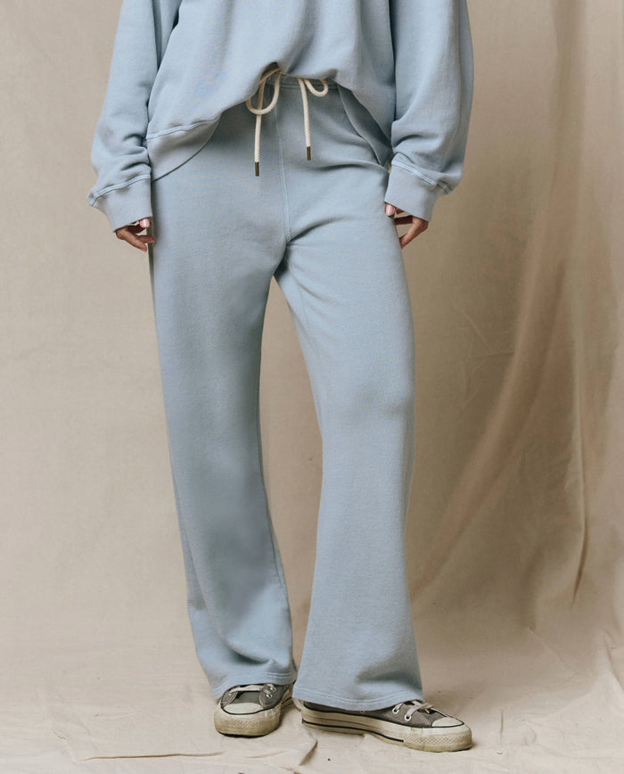 A person wears THE GREAT. SP26 SEASONAL CORE The Lap Sweatpant in Whisper Blue and a matching drawstring sweatshirt, standing on a beige fabric backdrop. Each piece is thoughtfully handcrafted in Los Angeles.