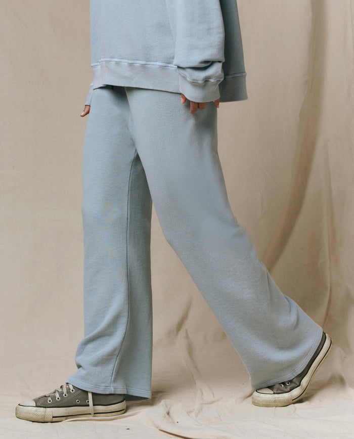 A person in wide leg sweatpants, an oversized sweatshirt, and gray sneakers stands against a beige background. The look highlights THE GREAT.’s Lap Sweatpant in Whisper Blue from the SP26 Seasonal Core collection for relaxed style.