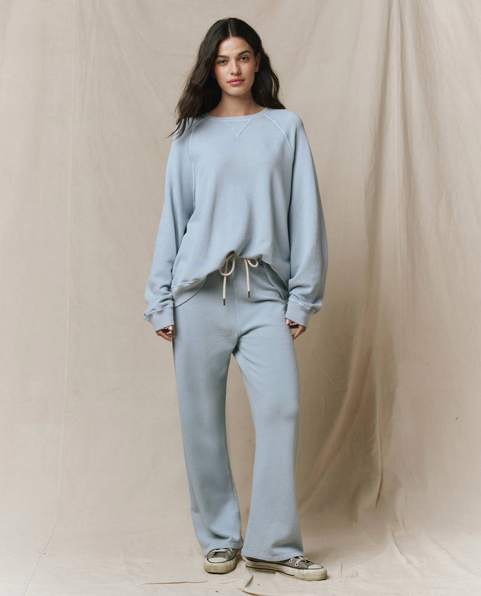 A woman stands against a beige fabric backdrop, wearing a loose light blue sweatshirt and THE GREAT. Lap Sweatpant in Whisper Blue from the SP26 SEASONAL CORE collection, styled with white sneakers.