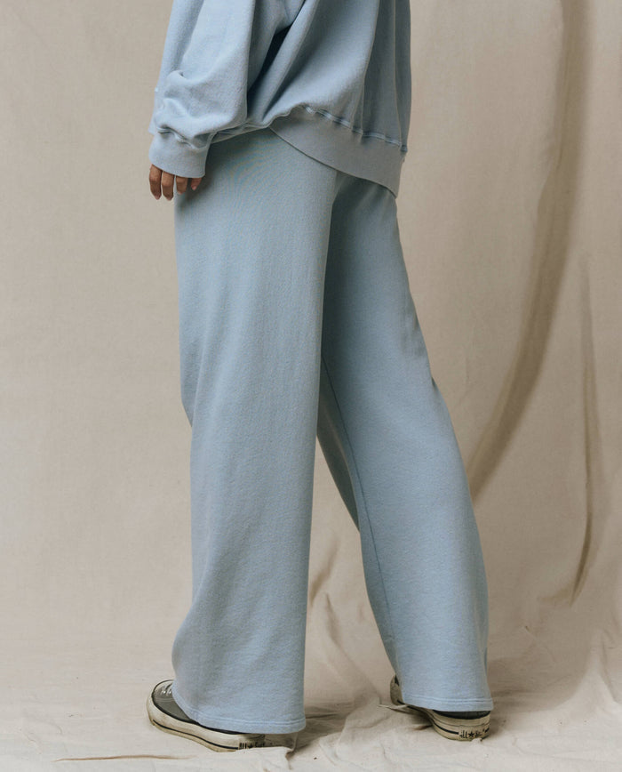 A person stands sideways on a neutral fabric backdrop wearing only the lower half, showcasing THE GREAT. SP26 SEASONAL CORE The Lap Sweatpant in Whisper Blue—loose, wide leg sweatpants with a handcrafted finish.