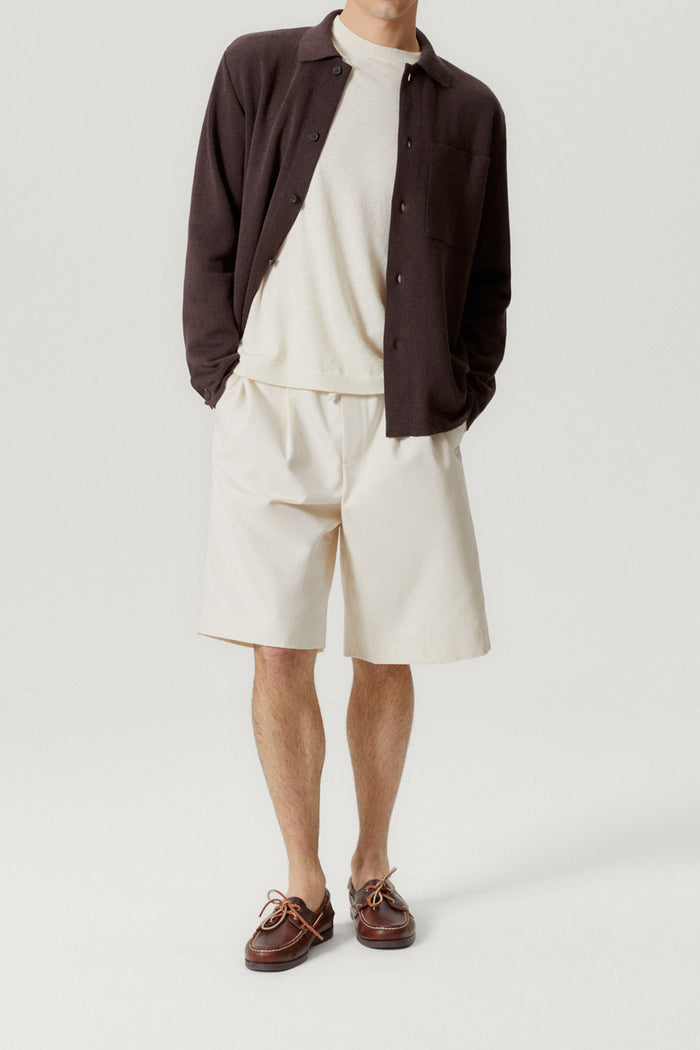Person wearing a brown overshirt, white sweater, beige shorts, and brown shoes on a light gray background