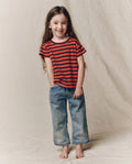 A young child with long hair stands barefoot on a neutral background, smiling with hands in pockets while wearing THE GREAT. F26 D1’s The Little Boxy Crew in Hot Red Naval Stripe and blue jeans.