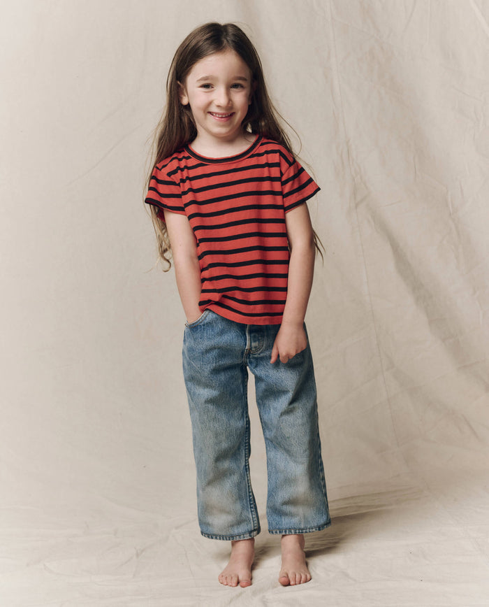 A young child with long hair stands barefoot on a neutral background, smiling with hands in pockets while wearing THE GREAT. F26 D1’s The Little Boxy Crew in Hot Red Naval Stripe and blue jeans.