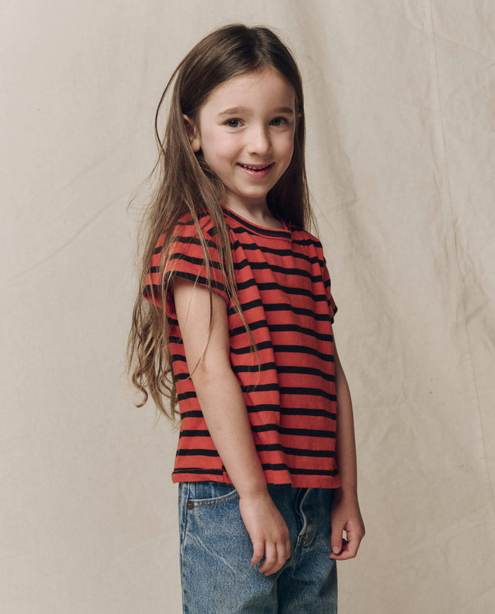 A young child with long brown hair, wearing THE GREAT. F26 D1 The Little Boxy Crew in Hot Red Naval Stripe and blue jeans, stands smiling in front of a plain beige background.