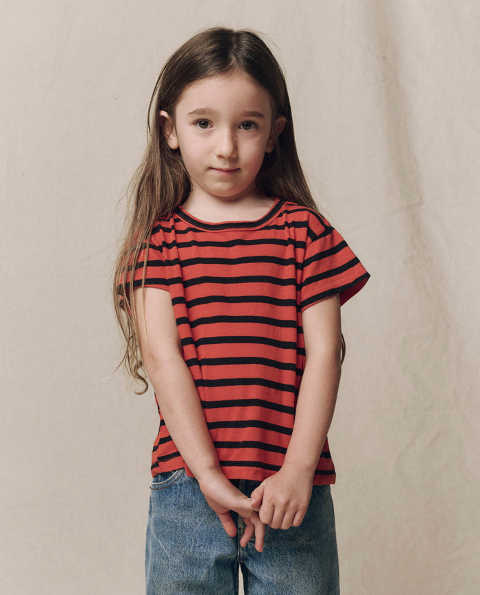 A young child with long brown hair wears THE GREAT.’s The Little Boxy Crew in Hot Red Naval Stripe, paired with blue jeans, standing against a neutral background with hands clasped.