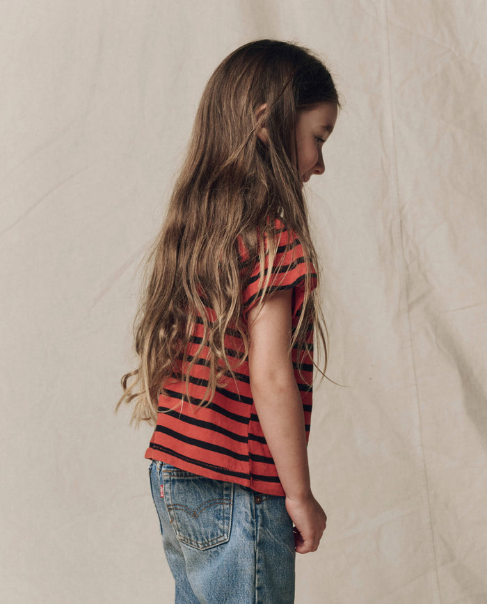 A child with long brown hair stands sideways against a beige background, wearing THE GREAT. F26 D1′s The Little Boxy Crew in Hot Red Naval Stripe and blue jeans.
