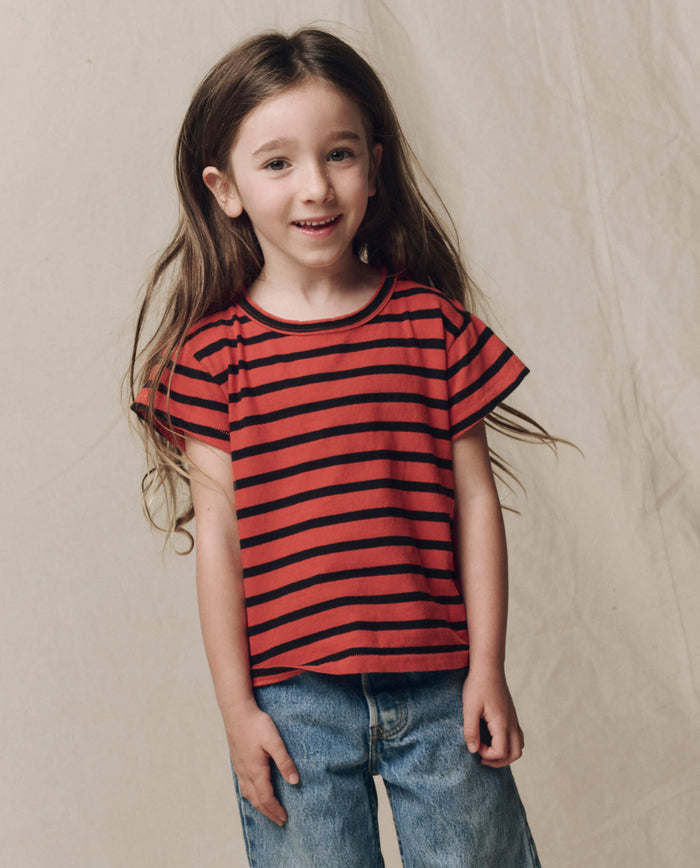 A young child with long brown hair wears THE GREAT. F26 D1 The Little Boxy Crew in Hot Red Naval Stripe and blue jeans, standing in front of a neutral background.
