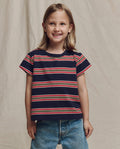 A young child smiles in front of a beige backdrop, wearing THE GREAT. SP26 LITTLES The Little Boxy Crew -- Scout Stripe and blue jeans.