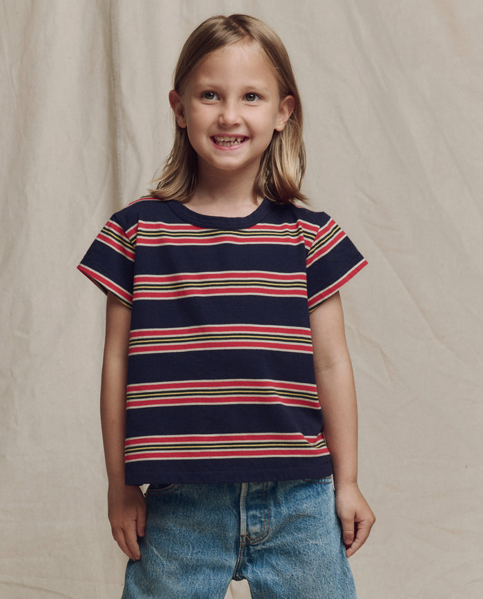 A young child smiles in front of a beige backdrop, wearing THE GREAT. SP26 LITTLES The Little Boxy Crew -- Scout Stripe and blue jeans.
