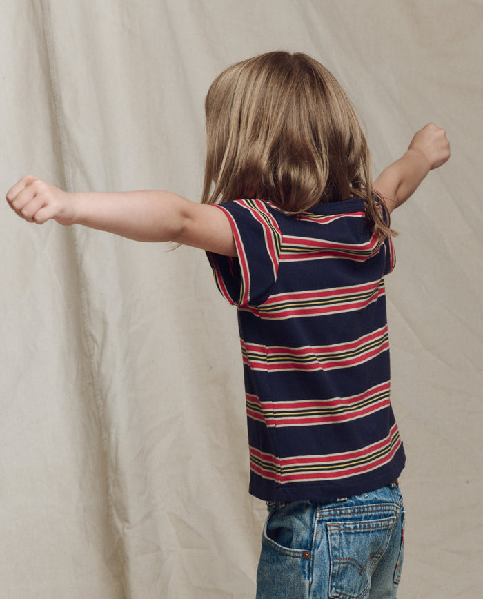 A child with long hair, wearing THE GREAT. SP26 LITTLES The Little Boxy Crew -- Scout Stripe and jeans, stands with arms outstretched facing away from the camera.