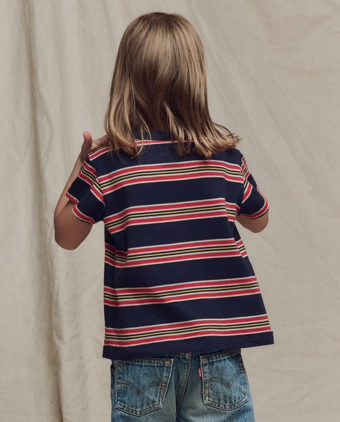 A child with long light brown hair faces away, wearing THE GREAT. SP26 LITTLES The Little Boxy Crew in Scout Stripe and blue jeans, standing against a plain beige background.