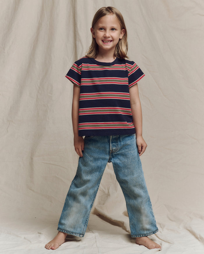A child with long hair smiles barefoot on a light fabric backdrop, wearing THE GREAT. SP26 LITTLES The Little Boxy Crew in Scout Stripe and loose blue jeans.