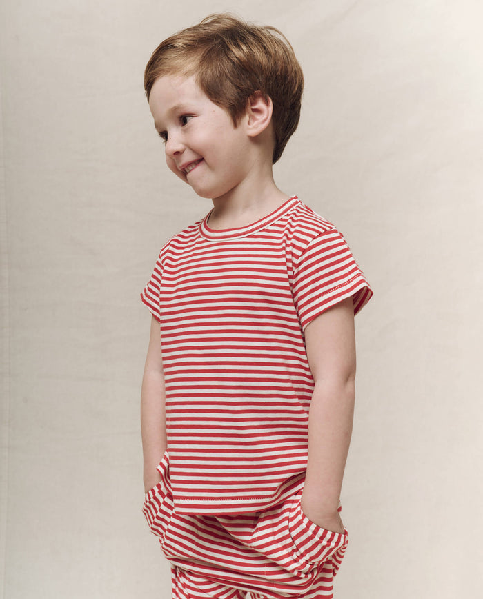 A young child smiles in a Sun Cherry Stripe Little Boxy Crew shirt and pants by THE GREAT. SP25 POLO LOUNGE LITTLE, set against a plain backdrop.