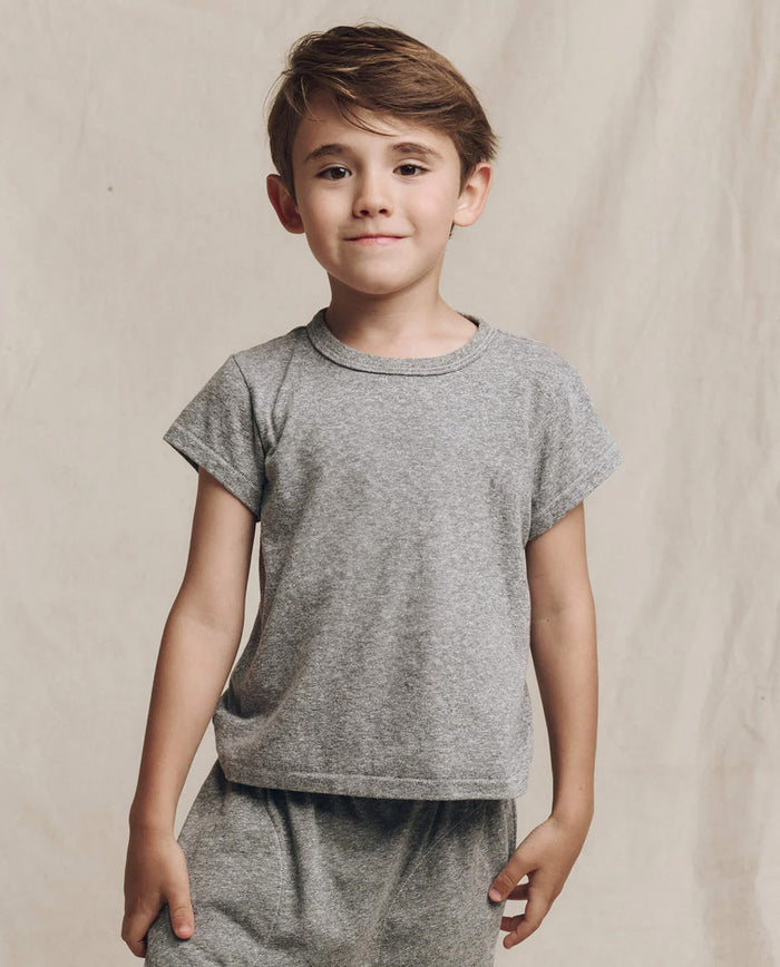 A child in a Heather Grey "The Little Boxy Crew" t-shirt by THE GREAT. LITTLE stands against a beige background, radiating a vintage vibe with its angled sleeves.