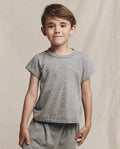 A child in a Heather Grey "The Little Boxy Crew" t-shirt by THE GREAT. LITTLE stands against a beige background, radiating a vintage vibe with its angled sleeves.
