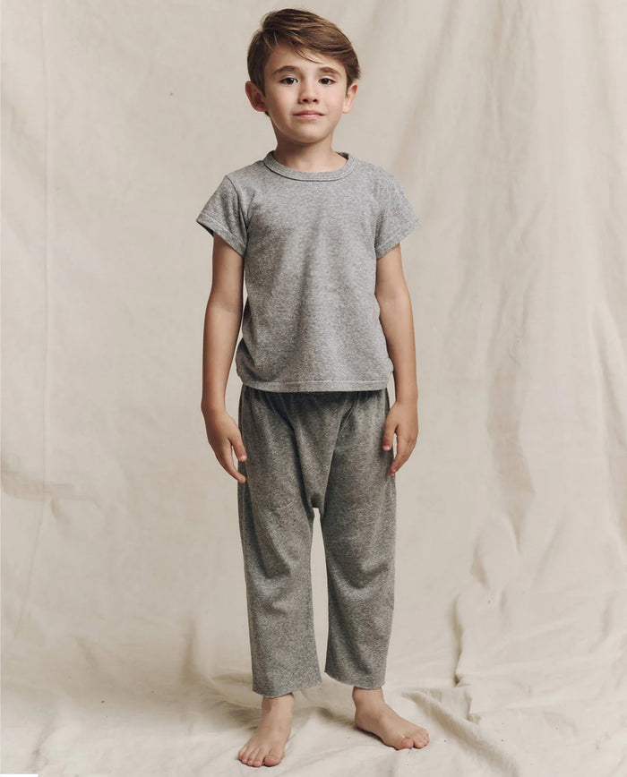 A young boy in "The Little Boxy Crew - Heather Grey" by THE GREAT. LITTLE stands barefoot on a textured, light-colored cloth backdrop, evoking a vintage feel.