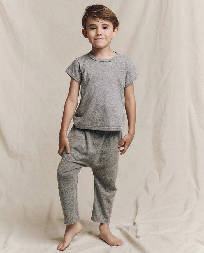 A child wearing The Little Boxy Crew in Heather Grey by THE GREAT. LITTLE, featuring angled sleeves for a vintage touch, stands barefoot against a neutral fabric backdrop.