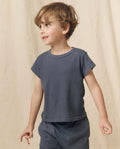 A young boy with light brown hair wears THE GREAT. LITTLE’s The Little Boxy Crew Solid in Washed Navy, a short-sleeve 100% cotton t-shirt paired with matching shorts, standing against a beige background for a subtle vintage feel.
