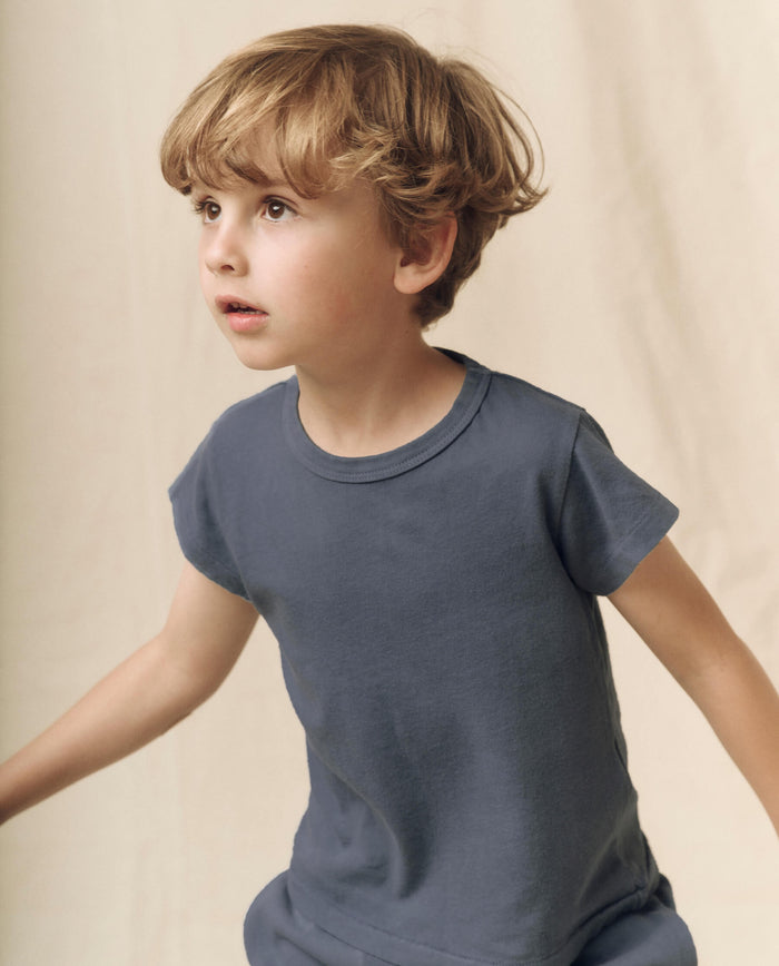 A young child with light brown hair wears The Little Boxy Crew in Washed Navy by THE GREAT. LITTLE—a short-sleeved, 100% cotton tee designed for a vintage feel—while looking slightly to the left against a plain, neutral background.