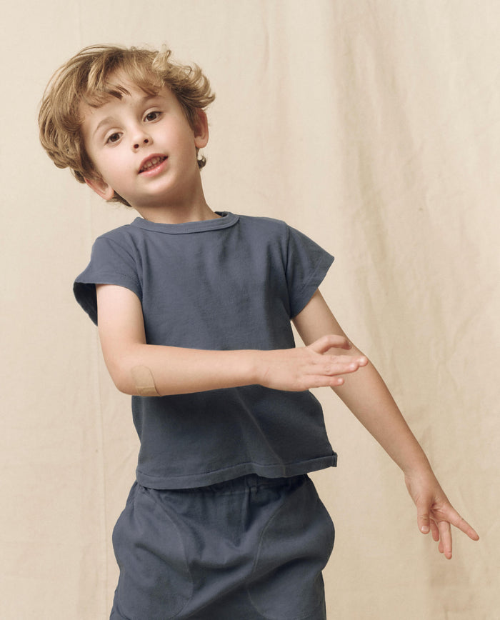 A young child with light brown hair wears THE GREAT. LITTLE’s The Little Boxy Crew in Solid Washed Navy with matching shorts, posing with one arm raised in front of a beige fabric backdrop for a vintage-inspired look.