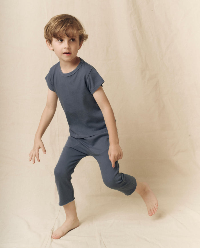 A young child with light brown hair wears THE GREAT. LITTLE’s The Little Boxy Crew in Solid -- Washed Navy, paired with matching pants, standing on one leg against a neutral-colored fabric backdrop for a subtle vintage feel.