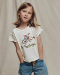 A child with shoulder-length hair wears THE GREAT. SP26 LITTLES' The Little Boxy Crew w/ Rider Graphic in Washed White and blue jeans, standing hands-in-pockets against a beige fabric backdrop.
