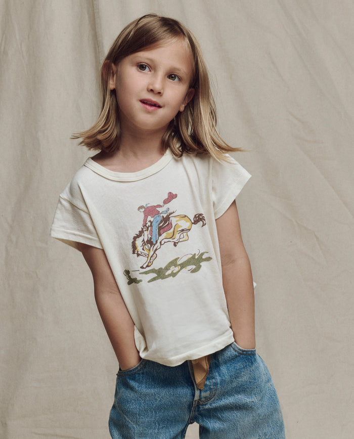 A child with shoulder-length hair wears THE GREAT. SP26 LITTLES' The Little Boxy Crew w/ Rider Graphic in Washed White and blue jeans, standing hands-in-pockets against a beige fabric backdrop.