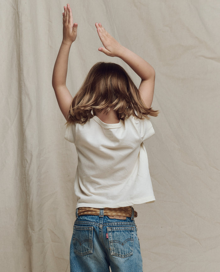 A child with shoulder-length hair wears THE GREAT. SP26 LITTLES The Little Boxy Crew w/ Rider Graphic in washed white, paired with blue jeans, standing with arms raised against a plain beige background.