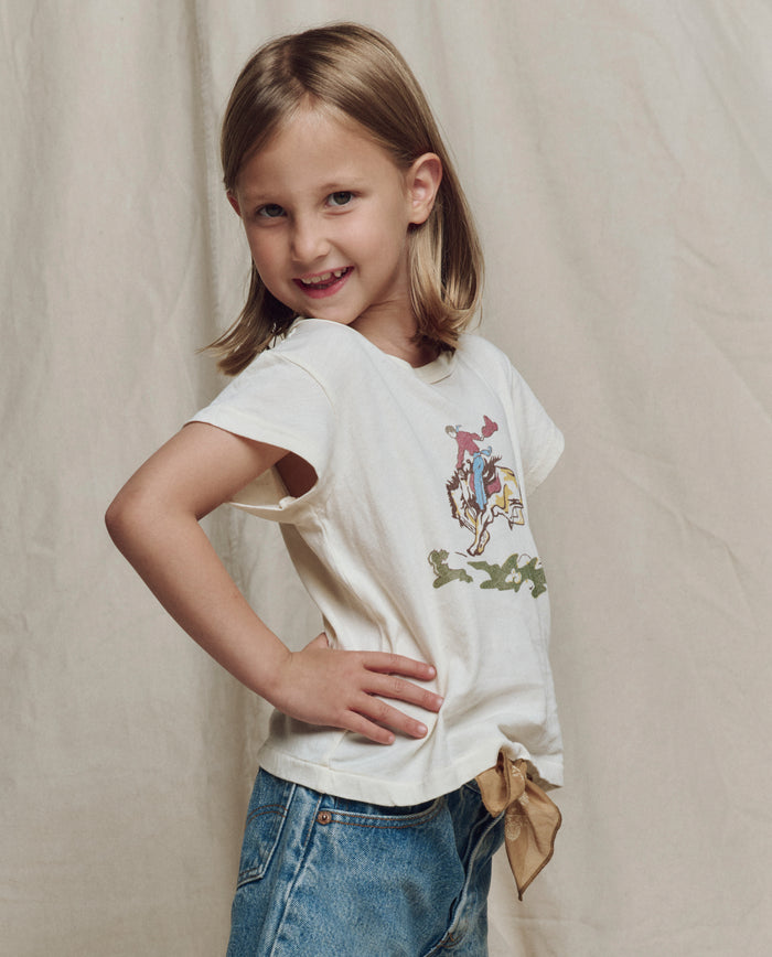 A young girl with light brown hair stands confidently in front of a beige background, wearing THE GREAT. SP26 LITTLES’ The Little Boxy Crew w/ Rider Graphic in washed white and blue jeans.