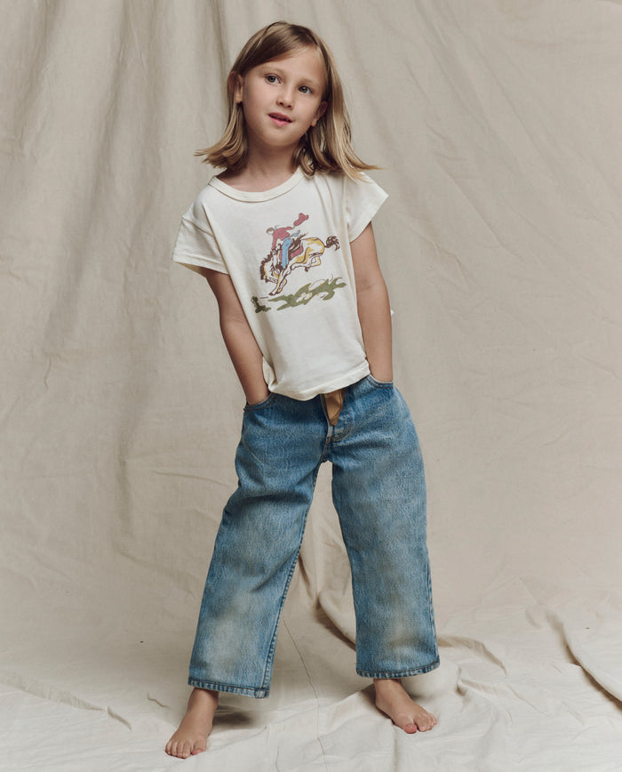 A young child stands barefoot on fabric, wearing THE GREAT. SP26 LITTLES The Little Boxy Crew w/ Rider Graphic in Washed White and loose blue jeans, hands in pockets.