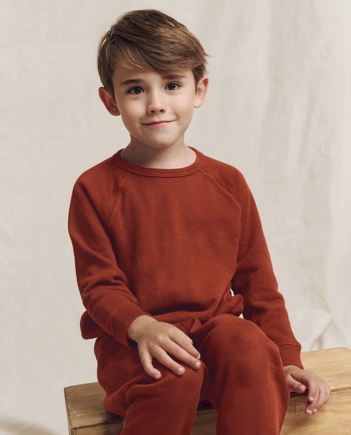 A young boy with brown hair wears THE GREAT. F25 LITTLES The Little College Sweatshirt in Solid Brick, paired with pants, as he sits on a wooden bench against a plain light background.