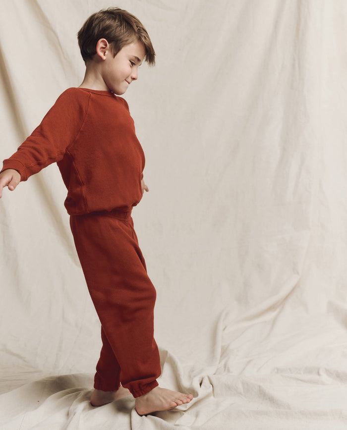A young child wears THE GREAT. F25 LITTLES The Little College Sweatshirt in Solid Brick, standing barefoot against a light fabric backdrop.