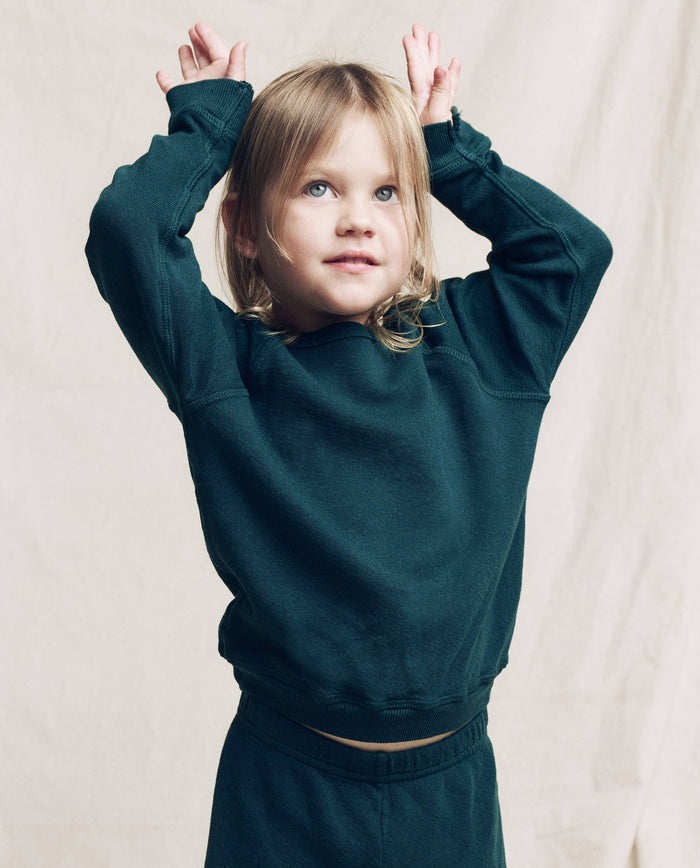 A young child wears THE GREAT. F25 LITTLES The Little College Sweatshirt, Solid -- Dark Slate, standing against a beige background with arms raised and hands near their head.