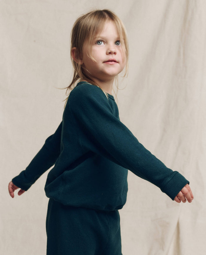 A young child with blonde hair wears THE GREAT. F25 LITTLES The Little College Sweatshirt, Solid in Dark Slate, paired with matching pants and stands with arms outstretched against a plain beige background.