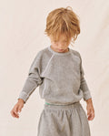 A young child with blond hair, wearing THE GREAT. FALL21 D2 LITTLE's The Little College Sweatshirt in Varsity Grey and soft polyester and cotton sweatpants, stands against a neutral background looking down.