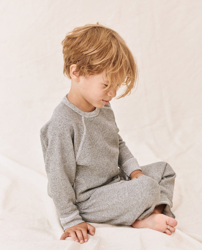 A young child with light brown hair sits cross-legged and looks down, wearing a cozy The Little College Sweatshirt in Varsity Grey from THE GREAT. FALL21 D2 LITTLE, along with matching pants. The background is plain and light-colored.
