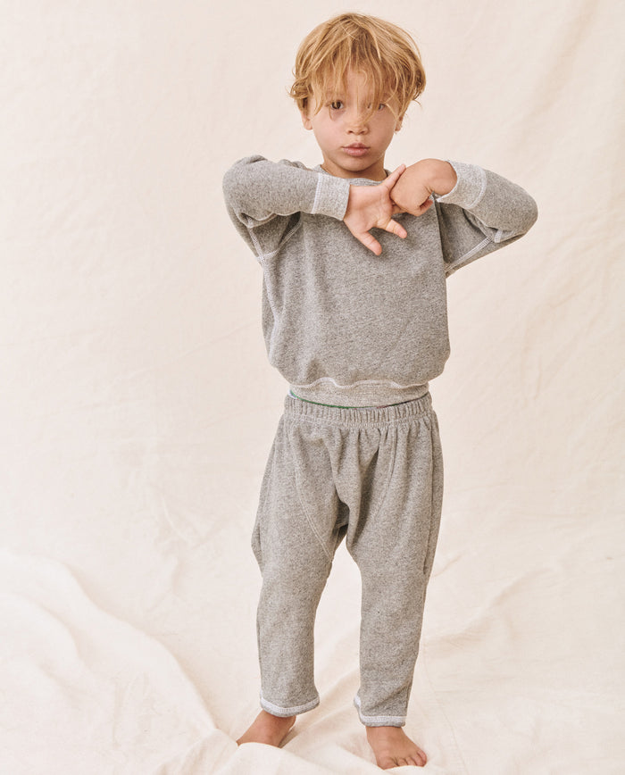 A young child with blond hair standing against a neutral background, wearing The Little College Sweatshirt in Varsity Grey from THE GREAT. FALL21 D2 LITTLE, with arms raised in a relaxed pose.