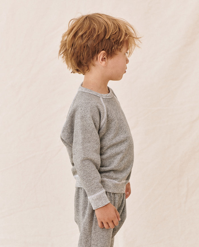 A young child with tousled hair stands in profile, wearing The Little College Sweatshirt in Varsity Grey by THE GREAT. FALL21 D2 LITTLE, paired with matching pants against a plain, light-colored background.